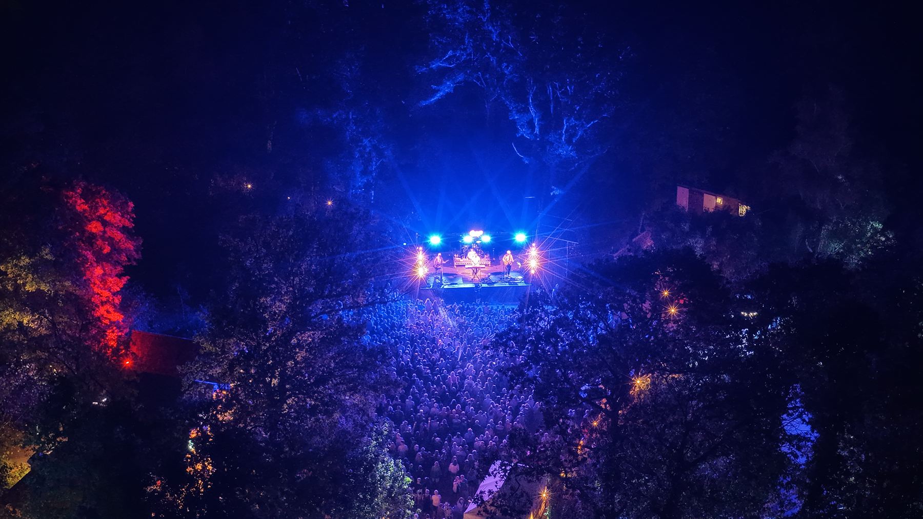 Night aerial photograph over a woodland festival stage, the crowd lit blue beneath the canopy