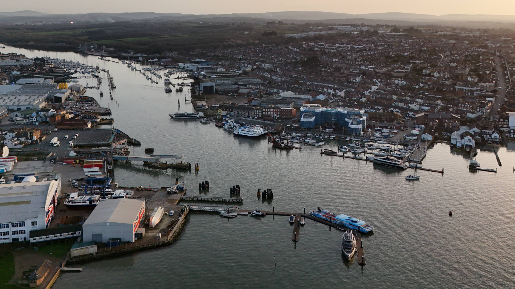 Aerial photograph over Cowes and the River Medina at dusk, showing marina pontoons, a boatyard, a ferry berth, industrial units and dense housing